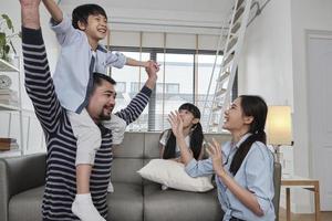 familia asiática tailandesa unida, papá juega y bromea con los niños y la mamá cargando y sosteniendo a su hijo sobre los hombros en la sala de estar de la casa, tiempos de ocio felices, fin de semana encantador, estilo de vida doméstico de bienestar. foto