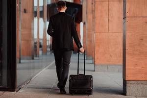 Going to airport terminal. Confident businessman traveler walking on city streets and pulling his suitcase drinking coffee and speaking on smartphone photo