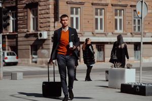 Going to airport terminal. Confident businessman traveler walking on city streets and pulling his suitcase drinking coffee and speaking on smartphone photo