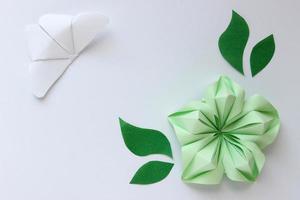 Origami white paper background with butterfly, green flower and leaves. With place for text. Origami composition. Paper craft photo