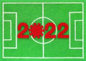 Red felt new year numbers 2022, zero in the form of a corona virus, placed on mini football field made of green felt, top view. Concept about soccer championship during covid pandemic. photo