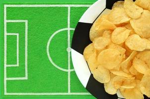 Football soccer party table with snacks. Salted potato chips on big plate painted like soccer ball, on dish mat background like small football field made of green felt. Top view, place for your text. photo