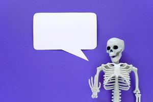 Skeleton on violet background with white blank paper rectangular speech bubble. Anatomical plastic model human skeleton with narration text balloon. Empty mockup dialog cloud. Purple Halloween. photo