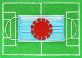 Red felt covid virus centered on blue medical mask placed on mini football field made of green felt, top view. Concept wear respirators and protect yourself from coronavirus on stadium soccer match. photo