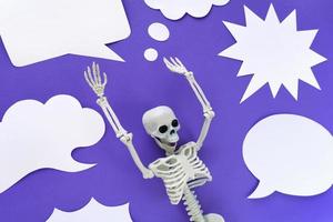 esqueleto sobre fondo violeta con un montón de burbujas de habla de papel blanco en blanco. esqueleto humano modelo plástico anatómico con las manos arriba y variedad de emociones. nubes de diálogo vacías. Víspera de Todos los Santos púrpura. foto