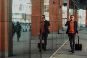 Going to airport terminal. Confident businessman traveler walking on city streets and pulling his suitcase drinking coffee and speaking on smartphone photo
