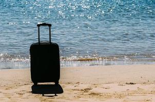 Black travel suitcase on sandy beach with turquoise sea background, summer holidays concept photo