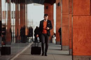 Going to airport terminal. Confident businessman traveler walking on city streets and pulling his suitcase drinking coffee and speaking on smartphone photo