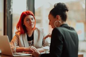 Two young business women sitting at table in cafe. Girl shows colleague information on laptop screen. Girl using smartphone, blogging. Teamwork, business meeting. photo