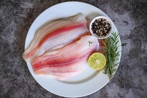 filete de pescado pangasius crudo fresco con hierbas y especias, lima limón y romero, pescado de carnita tilapia, bagre rayado, filete de pescado en un plato blanco con ingredientes para cocinar - vista superior foto