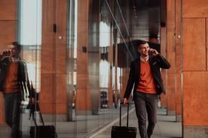 Going to airport terminal. Confident businessman traveler walking on city streets and pulling his suitcase drinking coffee and speaking on smartphone photo