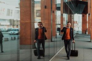 ir a la terminal del aeropuerto. viajero de negocios confiado caminando por las calles de la ciudad y tirando de su maleta bebiendo café y hablando en el teléfono inteligente foto