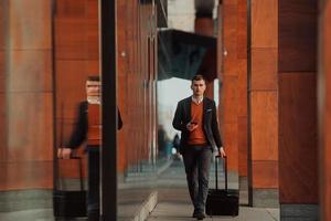 ir a la terminal del aeropuerto. viajero de negocios confiado caminando por las calles de la ciudad y tirando de su maleta bebiendo café y hablando en el teléfono inteligente foto