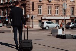 Going to airport terminal. Confident businessman traveler walking on city streets and pulling his suitcase drinking coffee and speaking on smartphone photo