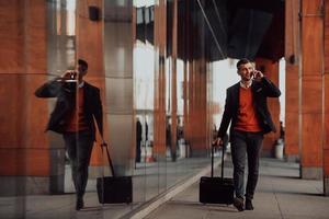 ir a la terminal del aeropuerto. viajero de negocios confiado caminando por las calles de la ciudad y tirando de su maleta bebiendo café y hablando en el teléfono inteligente foto