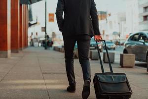 ir a la terminal del aeropuerto. viajero de negocios confiado caminando por las calles de la ciudad y tirando de su maleta bebiendo café y hablando en el teléfono inteligente foto