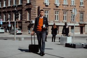 Going to airport terminal. Confident businessman traveler walking on city streets and pulling his suitcase drinking coffee and speaking on smartphone photo