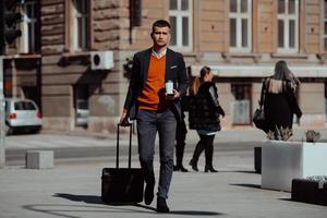 Going to airport terminal. Confident businessman traveler walking on city streets and pulling his suitcase drinking coffee and speaking on smartphone photo