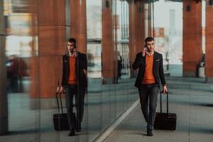 Going to airport terminal. Confident businessman traveler walking on city streets and pulling his suitcase drinking coffee and speaking on smartphone photo