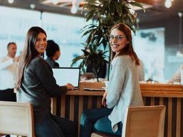 two young business women sitting at table in cafe. Girl shows colleague information on laptop screen. Girl using smartphone, blogging. Teamwork, business meeting.. photo