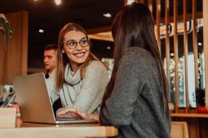 dos jóvenes mujeres de negocios sentadas a la mesa en el café. chica muestra información de colega en la pantalla del portátil. chica usando teléfono inteligente, blogueando. trabajo en equipo, reunión de negocios. foto