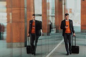 Going to airport terminal. Confident businessman traveler walking on city streets and pulling his suitcase drinking coffee and speaking on smartphone photo