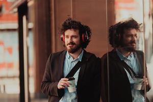 hombre de negocios de moda con maletín de cuero en el distrito financiero usando un teléfono inteligente mientras escucha canciones de audio con auriculares bebiendo café foto