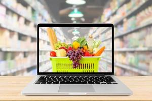 pasillo de supermercado fondo borroso con computadora portátil y cesta de compras en la mesa de madera concepto en línea de comestibles foto