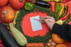 las verduras se disponen alrededor de una hoja de papel y un lápiz. espacio vacío para texto. mano femenina escribiendo una receta en un espacio en blanco vacío sobre un fondo rojo. foto