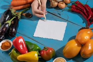 Vegetables are laid out around a sheet of paper and a pencil. Empty space for text. Vegetables, empty blank for recipe on a blue background. photo