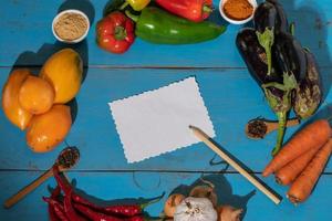 Vegetables are laid out around a sheet of paper and a pencil. Empty space for text. Vegetables, empty blank for recipe on a blue background. photo