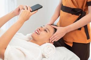 mujer joven feliz que usa el teléfono inteligente mientras está acostado en la mesa de masaje foto