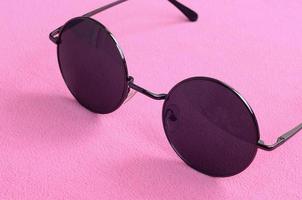 elegantes gafas de sol negras con gafas redondas se encuentran sobre una manta hecha de suave y esponjosa tela de lana rosa claro. imagen de fondo de moda en colores femeninos foto