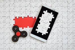 A modern big smartphone with a touch screen and a spinner lie on a white jigsaw puzzle in an assembled state with missing elements photo