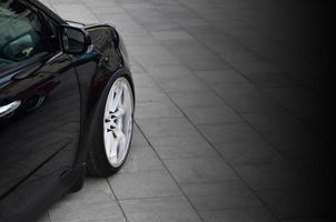 Diagonal view of a black glossy car with white wheels, which stands on a square of gray tiles photo