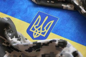 Ukrainian flag and coat of arms with fabric with texture of pixeled camouflage. Cloth with camo pattern in grey, brown and green pixel shapes with Ukrainian trident sign photo