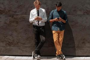 Casual multiethnic startup business men one of them is Indian using smartphone during break from work in front of grey wall outside photo
