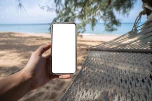 mano sosteniendo un móvil simulado con una pantalla blanca mientras está acostado en una hamaca columpiándose entre los árboles en la playa. foto