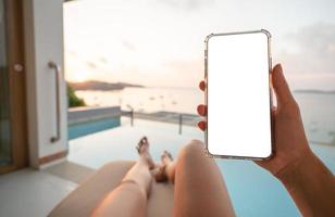 Woman holding mobile phone with blank screen while laying on sunbathing bed with beach view. photo