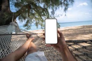 Hand holding mock up mobile with white sceen while laying on hammock swing between trees on the beach. photo