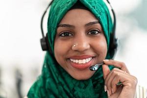 Afro Muslim female with green hijab scarf customer representative business woman with phone headset helping and supporting online with customer in modern call centre photo