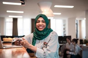 mujer de negocios musulmana africana que usa un hiyab verde usando un teléfono inteligente mientras trabaja en una computadora portátil en el área de relajación en la moderna oficina de inicio de planta abierta. foto