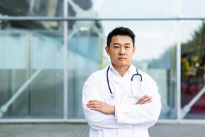 portrait of serious asian doctor with folded arms on background of modern clinic outdoors photo