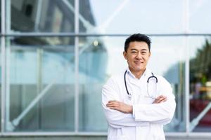 retrato de un alegre médico asiático sonriendo con las manos dobladas en el fondo de una clínica moderna foto