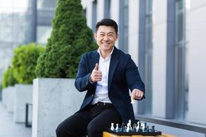 Cheerful Asian businessman playing chess in the yard smiling and looking forward to the camera photo