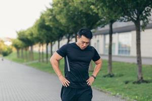 atleta asiático masculino, cansado después de un trote matutino, corre en el parque cerca del estadio foto