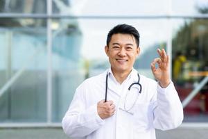 portrait of male cheerful asian doctor smiling and looking at camera showing ok hand sign photo