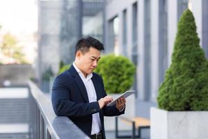 Serious asian boss reads news from tablet near modern office male businessman in business suit photo