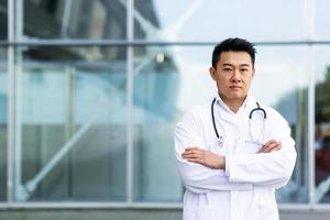 portrait of serious asian doctor with folded arms on background of modern clinic outdoors photo