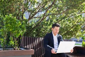 young asian business man or freelancer sitting on bench and working with laptop in city park photo